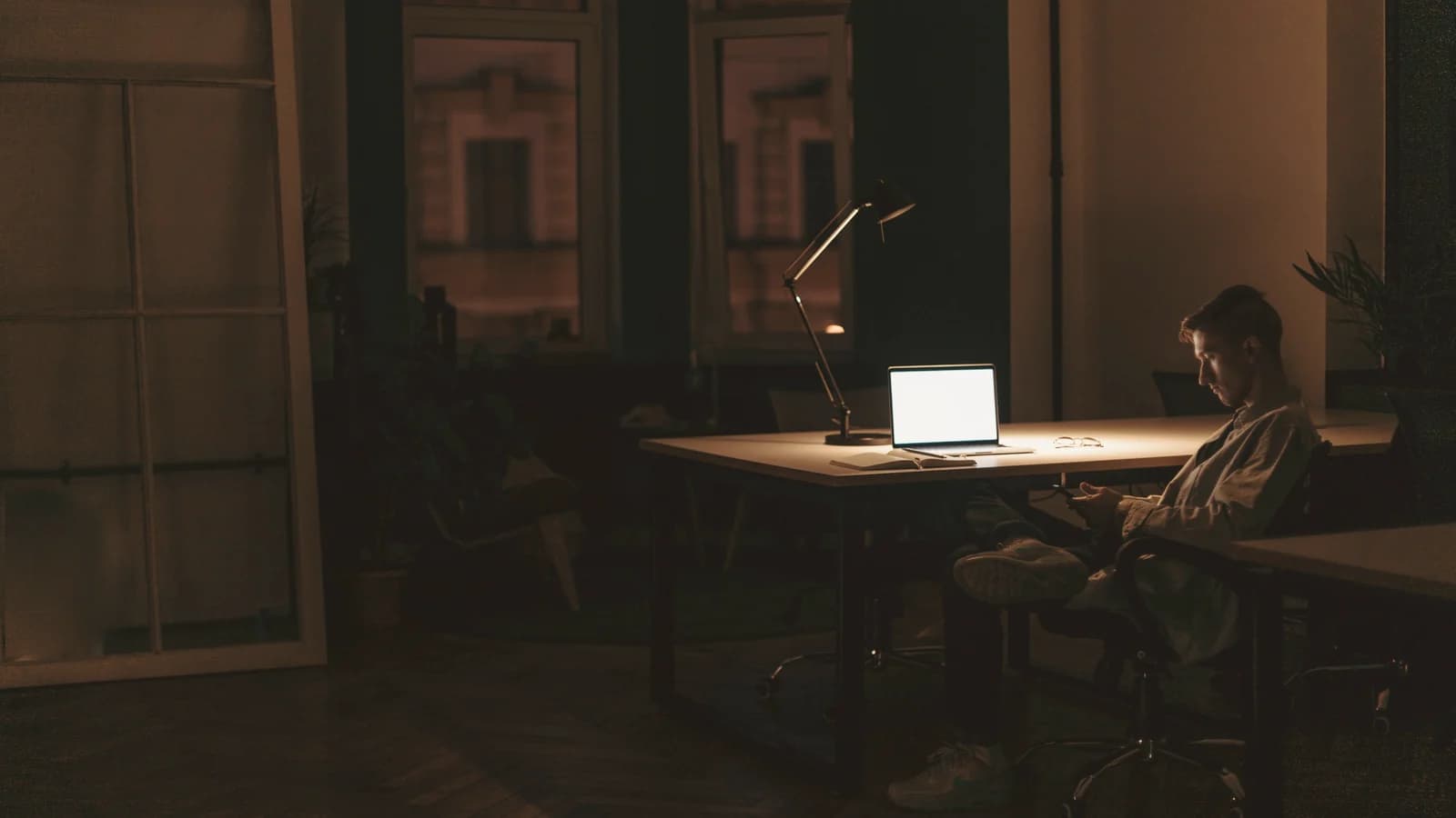 A man sits alone in a dark office in the evening, an open laptop and a desk lamp the only light
