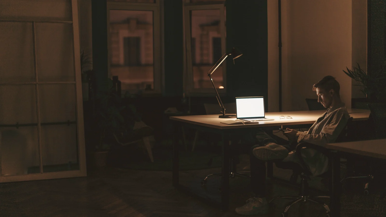 A man sits alone in a dark office in the evening, an open laptop and a desk lamp the only light
